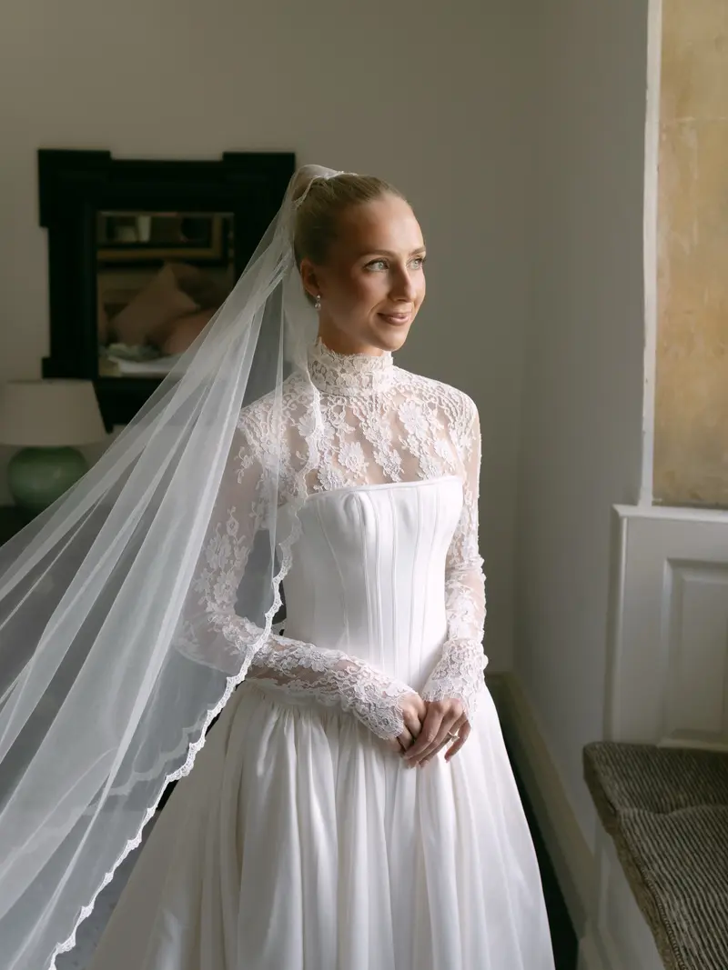 A bride in a lace gown and veil by a window